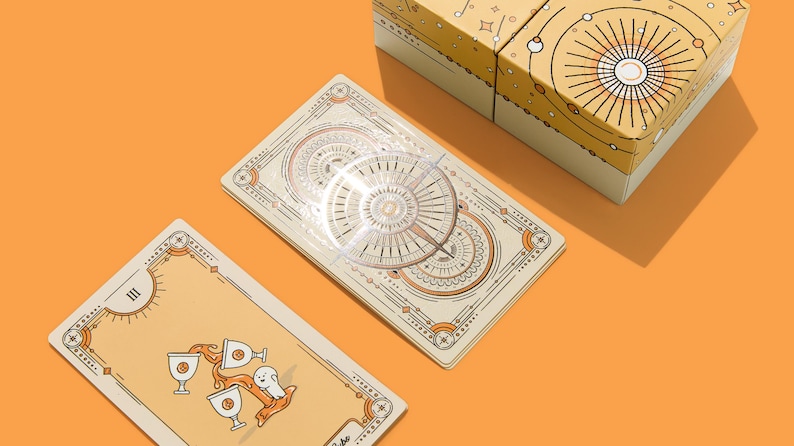 May include: A tarot card deck with a celestial design, featuring cream-colored cards with intricate illustrations in orange and brown. The deck is displayed with a matching box on an orange surface. One card shows three cups and a ghost.