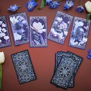 May include: A set of rectangular tarot cards featuring dark purple backgrounds with white floral illustrations and intricate silver-toned borders. Some cards display faces and helmets amidst flowers, while others show detailed circular geometric patterns.