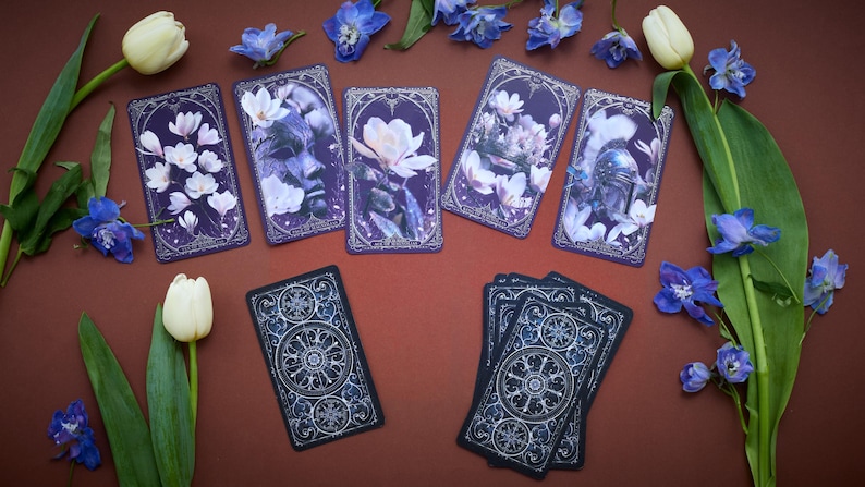 May include: A set of rectangular tarot cards featuring dark purple backgrounds with white floral illustrations and intricate silver-toned borders. Some cards display faces and helmets amidst flowers, while others show detailed circular geometric patterns.