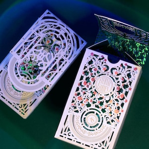 May include: Two rectangular playing card boxes featuring intricate white laser-cut floral overlays. The right box is partially open, revealing a shimmering green holographic interior. The text Eternal Garden is printed on the front of the right box.
