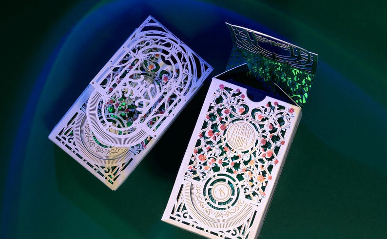 May include: Two rectangular playing card boxes featuring intricate white laser-cut floral overlays. The right box is partially open, revealing a shimmering green holographic interior. The text Eternal Garden is printed on the front of the right box.