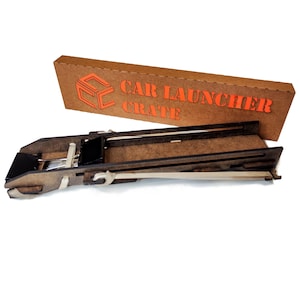 Könnte beinhalten: Ein Holz-Autostarter mit Gummiband und einem braunen Karton mit dem Text "CAR LAUNCHER CRATE".