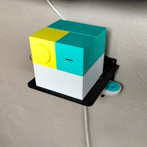 May include: A white, yellow, and teal cube-shaped device with a round yellow button and a black and white patterned surface. The device is mounted on a black shelf with a white cord.