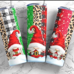May include: Three stainless steel tumblers with festive Christmas gnome designs. Each tumbler features a different pattern: black and white buffalo plaid, green glitter, and red glitter. The gnomes wear red hats and have white beards.