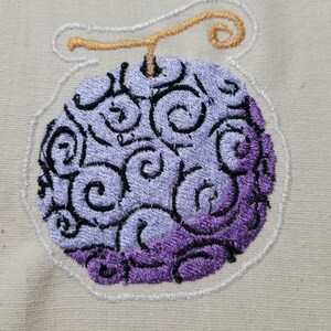 May include: Embroidered patch featuring a stylized, purple and lavender fruit with black swirl details. The fruit has a gold stem and is outlined in white thread. The patch is on a beige fabric background.