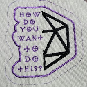 May include: Embroidered patch with the text "HOW DO YOU WANT TO DO THIS?" in purple thread. A black geometric design is on the right side. The patch is on a light beige fabric.