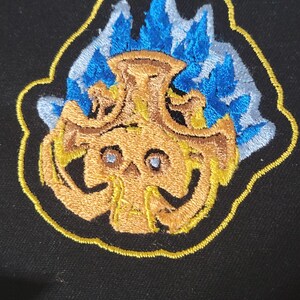 May include: Embroidered patch featuring a stylized skull with blue flames emanating from the top. The skull is primarily gold with blue accents, outlined in yellow, and set against a black background. The patch has a detailed, textured appearance.