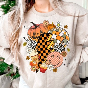 May include: A beige sweatshirt with a graphic design featuring a pumpkin, a disco ball, a chequered lightning bolt, a smiley face, a pumpkin spice latte, a slice of pumpkin pie, and a cupcake. The design is in shades of orange, yellow, brown, and white.