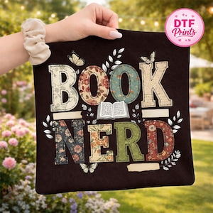 May include: A dark brown tote bag with the words "BOOK NERD" in a colorful, floral design. The letters are outlined in white with a stitched effect, and butterflies and leaves are scattered around the text.