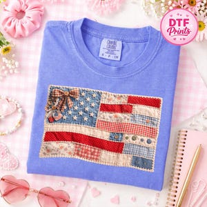 May include: A light blue t-shirt featuring a patchwork American flag design. The flag incorporates various patterned fabrics in red, white, and blue, with a bow and embellishments at the top. The shirt has a crew neck and the tag reads "Comfort Colors".
