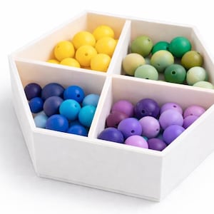 May include: A white, geometric wooden organizer filled with colorful beads. The beads are sorted into four sections, with yellow, green, blue, and purple beads. The beads are round and have a matte finish.