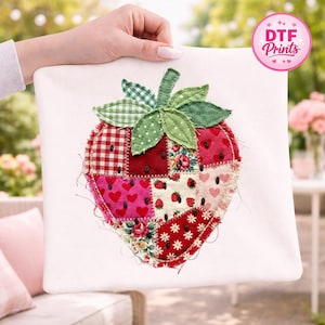 May include: A white pillow with a large, patchwork strawberry design. The strawberry is composed of various fabric patterns, including red gingham, floral prints, and heart motifs. Green leaves and a stem top the strawberry. The pillow is held up by a person.