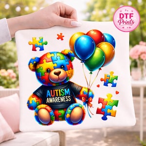May include: A white pillow featuring a colorful puzzle-piece bear holding balloons. The bear wears a black shirt with "AUTISM AWARENESS" printed on it. Additional puzzle pieces and a flower accent the design. The image is held by a person.