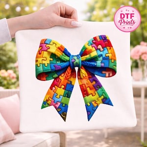 May include: A white canvas featuring a colorful bow design made of puzzle pieces. The bow is a vibrant mix of red, orange, yellow, green, blue, and purple. The image also includes a pink and white circular logo that says "DTF Prints".