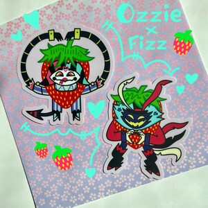 May include: Two cartoon stickers of strawberry characters with green hair and red and black clothing. The stickers are on a pink background with white flowers. The text "Ozzie x Fizz" is written in green and blue on the top of the image.