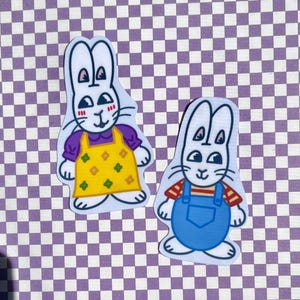 May include: Two white rabbit stickers with different outfits. One rabbit is wearing a purple shirt and a yellow dress with green and yellow flowers. The other rabbit is wearing a red and white striped shirt and blue overalls.