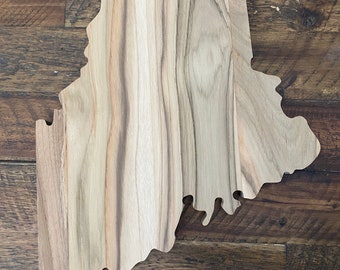 Puede incluir: Una tabla de cortar de madera con forma del estado de Maine. La tabla tiene un acabado de madera natural y muestra la veta de la madera.