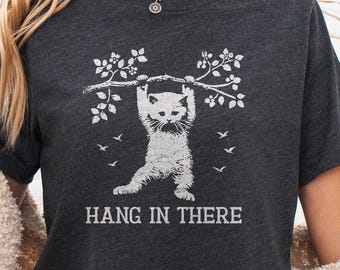 May include: A dark heather grey t-shirt featuring a white graphic of a kitten hanging from a tree branch with the text HANG IN THERE printed below. The design includes small leaves and birds surrounding the kitten on the branch.