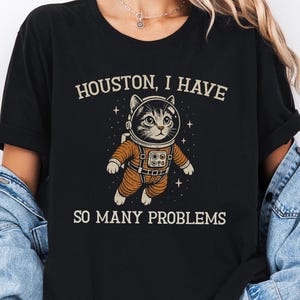 Puede incluir: Camiseta negra jaspeada con un gato de dibujos animados con traje de astronauta y el texto "Houston, I have so many problems." El gato lleva un traje espacial naranja, rodeado de estrellas. Una divertida camiseta gráfica con temática espacial.