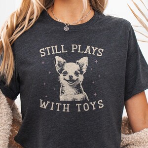 以下が含まれることがあります： 笑顔のチワワのイラストと「STILL PLAYS WITH TOYS」の文字が入ったダークヘザーグレーのTシャツ。カジュアルウェアです。