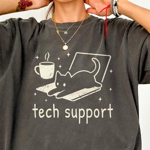 May include: A charcoal gray short-sleeve t-shirt featuring a white graphic of a cat lying on a laptop next to a coffee mug. The text "tech support" is printed below the graphic, surrounded by small star icons. The fabric has a washed-out finish.