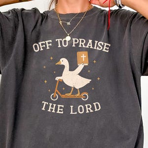 May include: A charcoal gray short-sleeve t-shirt featuring a graphic of a white goose riding a scooter and holding a brown book with a cross on the cover. The text reads OFF TO PRAISE THE LORD in a cream-colored font, surrounded by small star icons.