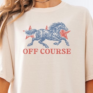Pode incluir: Camiseta cor marfim com um gráfico de cavalo azul desgastado, estrelas vermelhas e o texto "OFF COURSE" em vermelho. A camiseta tem mangas curtas e um corte folgado.