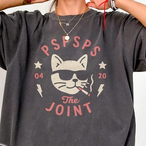 May include: Dark gray t-shirt with a graphic of a cat wearing sunglasses and smoking a cigarette. The design includes the text "PSPSPS" and "The Joint" in a pinkish-red color, along with stars, lightning bolts, and the numbers "04" and "20".