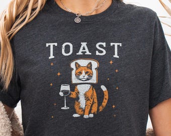 May include: A dark heather grey t-shirt featuring a graphic of an orange tabby cat with its head inside a slice of bread. The cat holds a wine glass, and the word TOAST is printed in white above the cat, surrounded by small star icons.