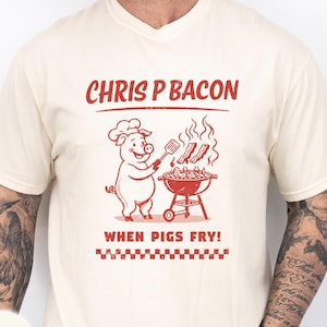 May include: Ivory-colored t-shirt with a red graphic of a pig chef grilling bacon. The text "CHRIS P BACON" is above the pig, and "WHEN PIGS FRY!" is below. A red and white checkered pattern is at the bottom.