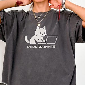 May include: Dark grey t-shirt featuring a pixelated cat working on a laptop. The text "PURRGRAMMER" is printed below the cat graphic in white. The shirt has a relaxed fit and short sleeves.