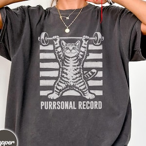 May include: Dark gray t-shirt featuring a gray and white illustration of a cat lifting a barbell. The cat is standing in front of white horizontal stripes. The text "PURRSONAL RECORD" is below the cat.