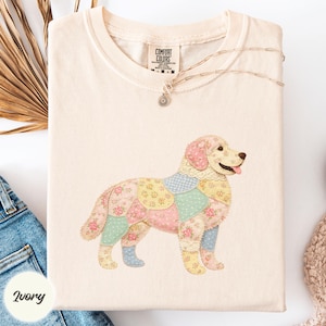 Puede incluir: Camiseta color marfil con un diseño de perro patchwork en patrones florales y cuadros pastel. El perro tiene una lengua rosa y un cuello de encaje. La camiseta tiene una etiqueta Comfort Colors y un collar plateado.
