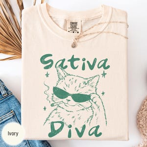 Può includere: Una t-shirt a maniche corte color avorio con una grafica verde scuro di un gatto che indossa occhiali da sole e fuma. Il testo Sativa Diva è stampato in verde scuro sopra e sotto l'illustrazione del gatto. La maglia ha un'etichetta Comfort Colors.