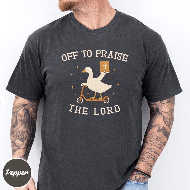 May include: A charcoal gray short-sleeve t-shirt featuring a graphic of a white goose riding a scooter and holding a brown book with a cross on the cover. The text "OFF TO PRAISE THE LORD" is printed in cream-colored letters above and below the goose.