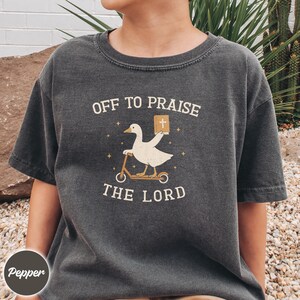 May include: A charcoal gray short-sleeve t-shirt featuring a graphic of a white goose riding a scooter and holding a book with a cross on the cover. The text reads OFF TO PRAISE THE LORD, surrounded by small star icons in a light cream color.