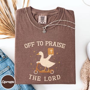 May include: A brown short-sleeve t-shirt featuring a graphic of a white goose riding a scooter and holding a book with a cross on the cover. The text reads OFF TO PRAISE THE LORD. The shirt has a Comfort Colors label at the inner neckline.