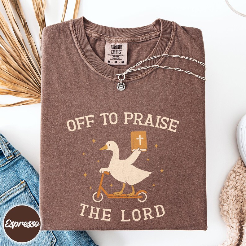May include: A brown short-sleeve t-shirt featuring a graphic of a white goose riding a scooter and holding a book with a cross on the cover. The text reads OFF TO PRAISE THE LORD. The shirt has a Comfort Colors label at the inner neckline.