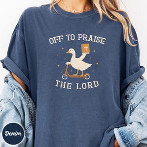 May include: A denim blue t-shirt featuring a graphic of a white goose riding a scooter and holding a book with a cross on the cover. The text reads OFF TO PRAISE THE LORD, surrounded by small star icons. The shirt has a crew neckline and short sleeves.