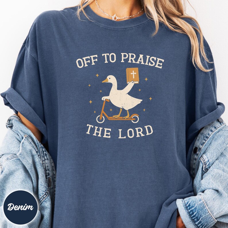 May include: A denim blue t-shirt featuring a graphic of a white goose riding a scooter and holding a book with a cross on the cover. The text reads OFF TO PRAISE THE LORD, surrounded by small star icons. The shirt has a crew neckline and short sleeves.