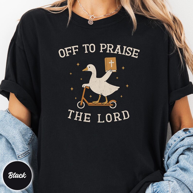 May include: A black t-shirt featuring a graphic of a white goose riding a kick scooter and holding a brown book with a white cross on the cover. The text "OFF TO PRAISE THE LORD" is printed in a cream-colored font above and below the goose illustration.