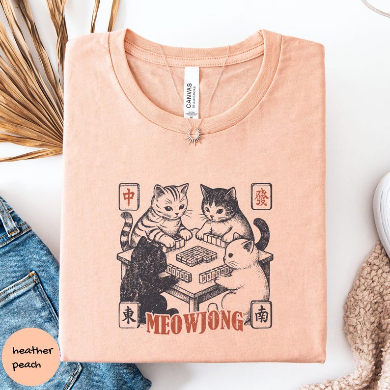 May include: A heather peach t-shirt featuring a black ink graphic of four cats playing a game of mahjong at a table. The design includes Chinese characters in four corners and the text MEOWJONG in a reddish-brown font at the bottom center.