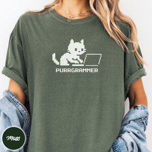 May include: A dark olive green t-shirt featuring a white pixel art graphic of a cat sitting at a laptop computer. Below the cat, the text PURRGRAMMER is printed in a matching white pixelated font. The shirt has a crew neckline and short sleeves.