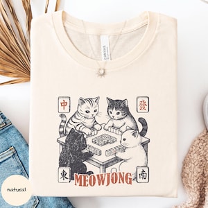 May include: A natural-colored t-shirt featuring a graphic print of four cats playing mahjong around a table. The design includes Chinese characters in red squares at the corners and the text MEOWJONG in red letters at the bottom of the illustration.
