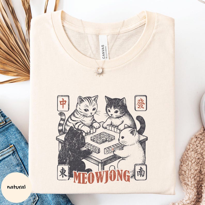 May include: A natural-colored t-shirt featuring a graphic print of four cats playing mahjong around a table. The design includes Chinese characters in red squares at the corners and the text MEOWJONG in red letters at the bottom of the illustration.