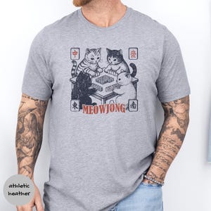 May include: An athletic heather gray short-sleeve t-shirt featuring a graphic of four cats playing mahjong at a table. The design includes the text MEOWJONG in red and various Chinese characters in square frames surrounding the illustration.