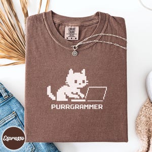 May include: A brown short-sleeve t-shirt featuring a light-colored pixel art graphic of a cat using a laptop with the text "PURRGRAMMER" printed below. The shirt has a Comfort Colors brand label at the inner neckline and a silver chain necklace.