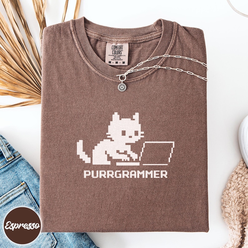 May include: A brown short-sleeve t-shirt featuring a light-colored pixel art graphic of a cat using a laptop with the text "PURRGRAMMER" printed below. The shirt has a Comfort Colors brand label at the inner neckline and a silver chain necklace.