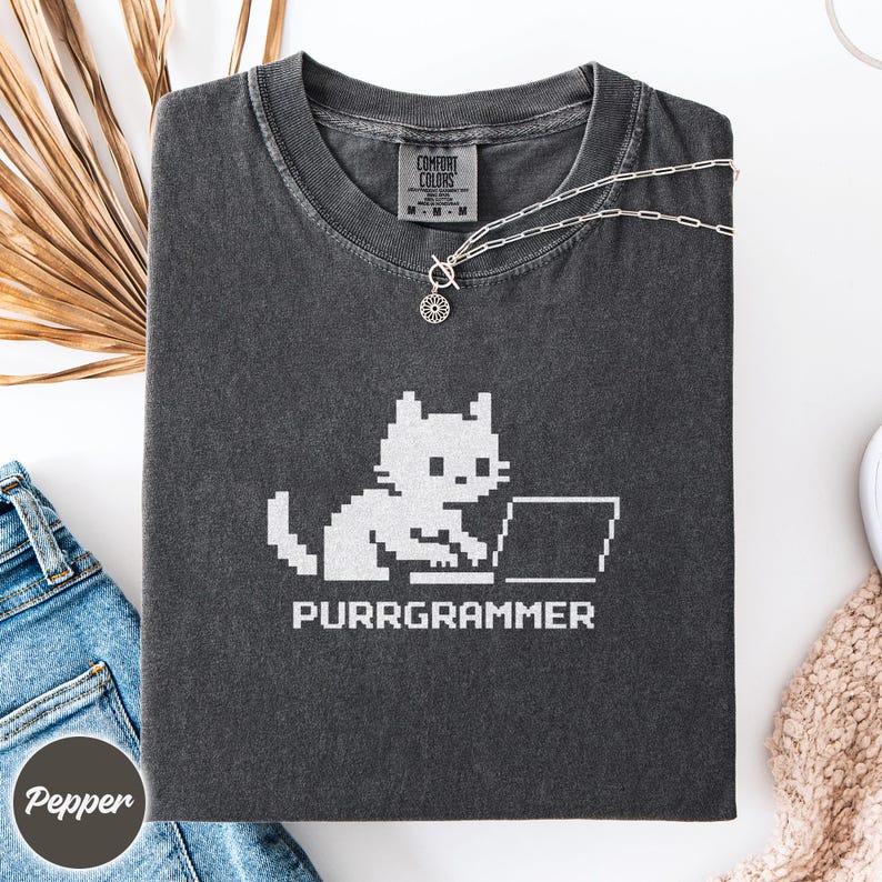 May include: A charcoal gray short-sleeve t-shirt featuring a white pixelated graphic of a cat sitting at a laptop with the text PURRGRAMMER below it. The shirt has a Comfort Colors brand label at the inner neckline and a crew neck collar.