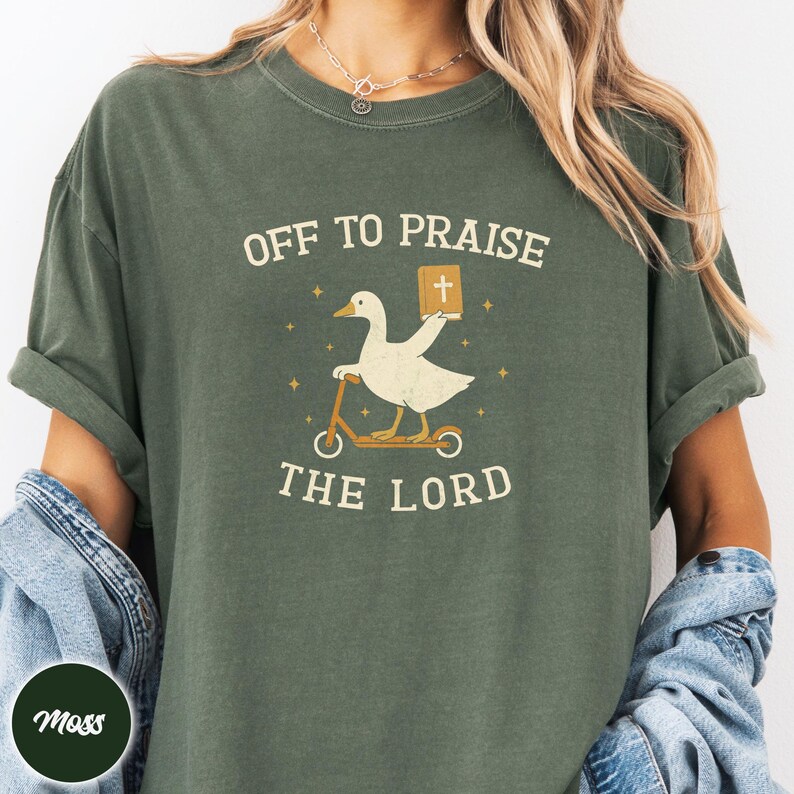 May include: A moss green t-shirt featuring a graphic of a white goose riding a scooter while holding a book with a cross on the cover. The text above the goose reads OFF TO PRAISE and the text below the goose reads THE LORD, surrounded by small stars.